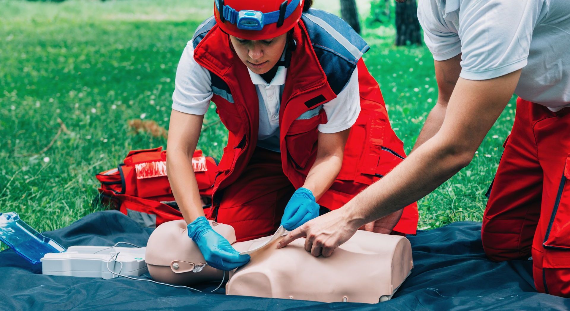Two people in red vests practice CPR on a mannequin in a park.