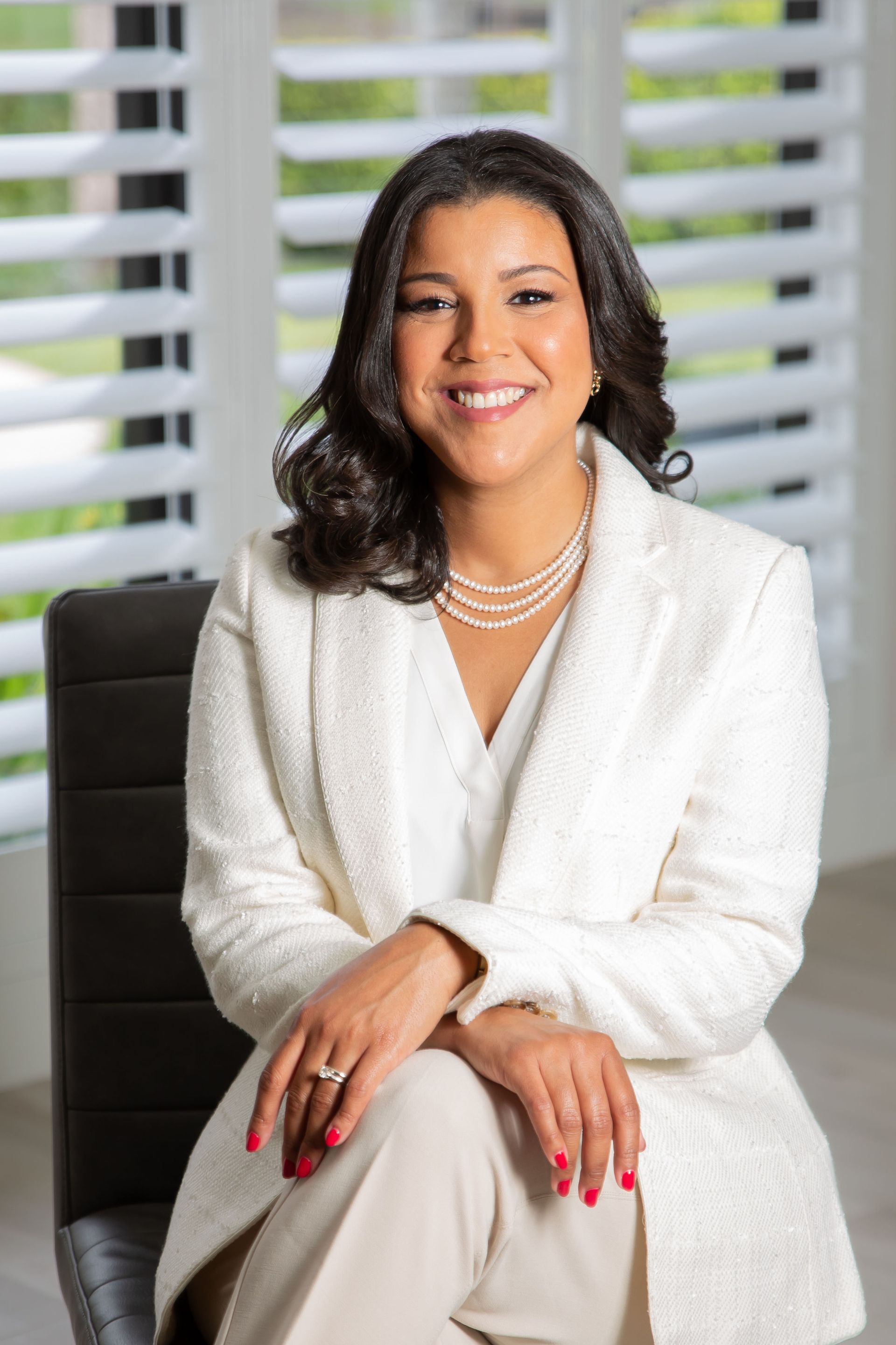 A woman in a white jacket and pearls is sitting in a chair.