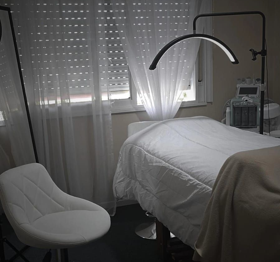 Una sala de tratamiento con una camilla de masaje, una lámpara estética curva y una silla blanca 