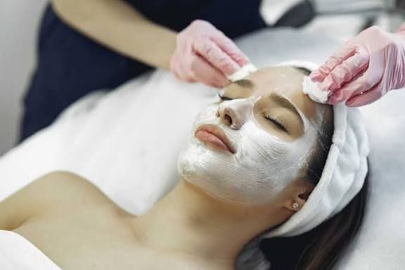 Una esteticista con guantes rosas limpia el rostro de una clienta cubierta con una mascarilla blanca