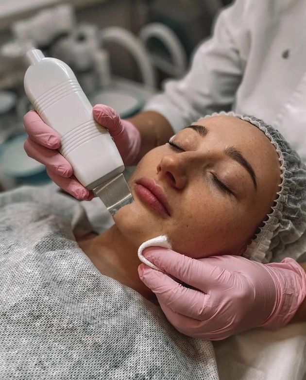 Una esteticista con guantes rosas limpia el rostro de una clienta cubierta con una mascarilla blanca