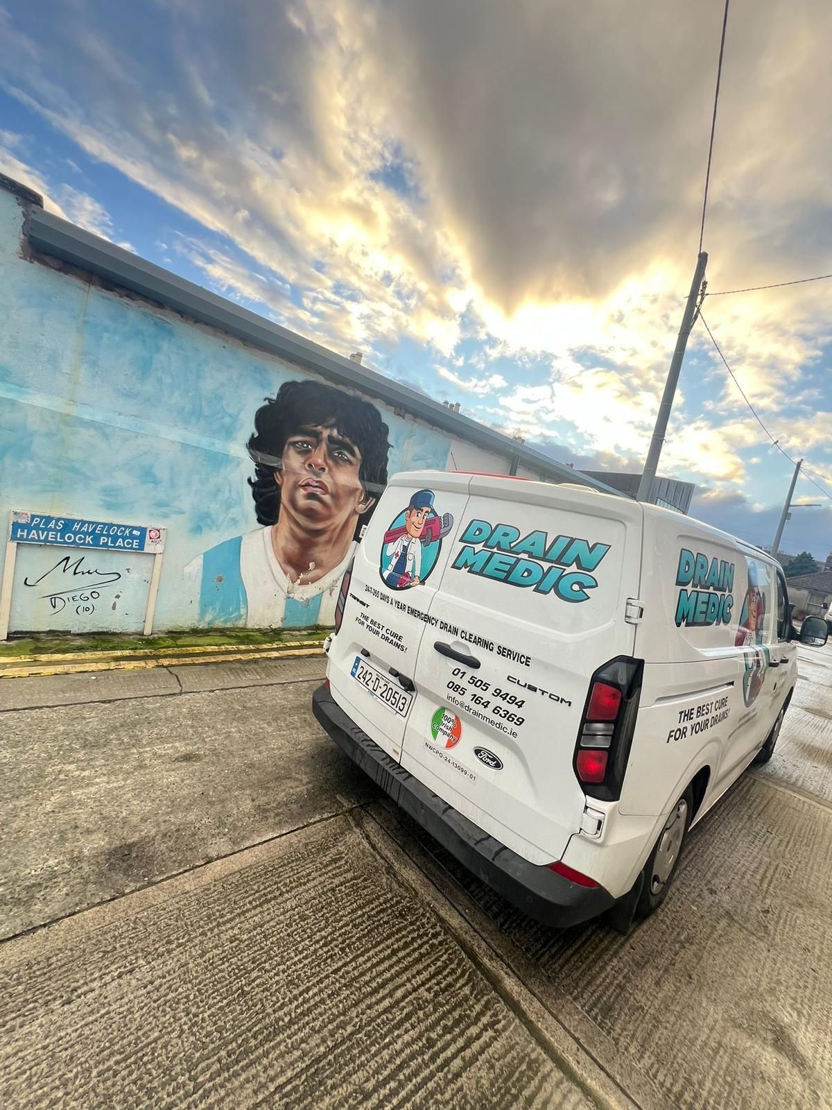 White van parked beside a mural-painted wall under a cloudy sunset sky