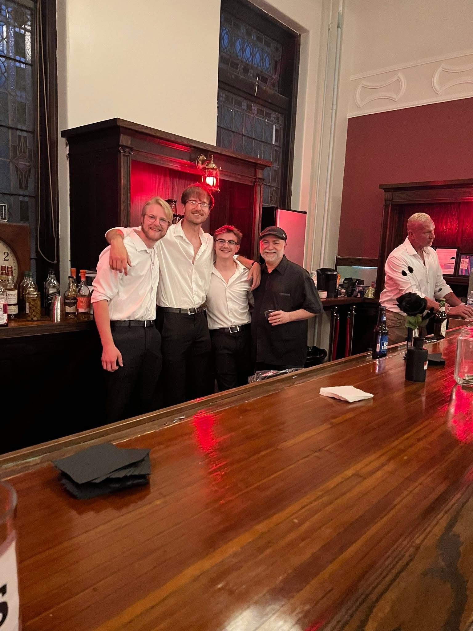 Four bartenders behind a wooden bar. Two people have their arms around each other. Dim lighting, red backdrop.