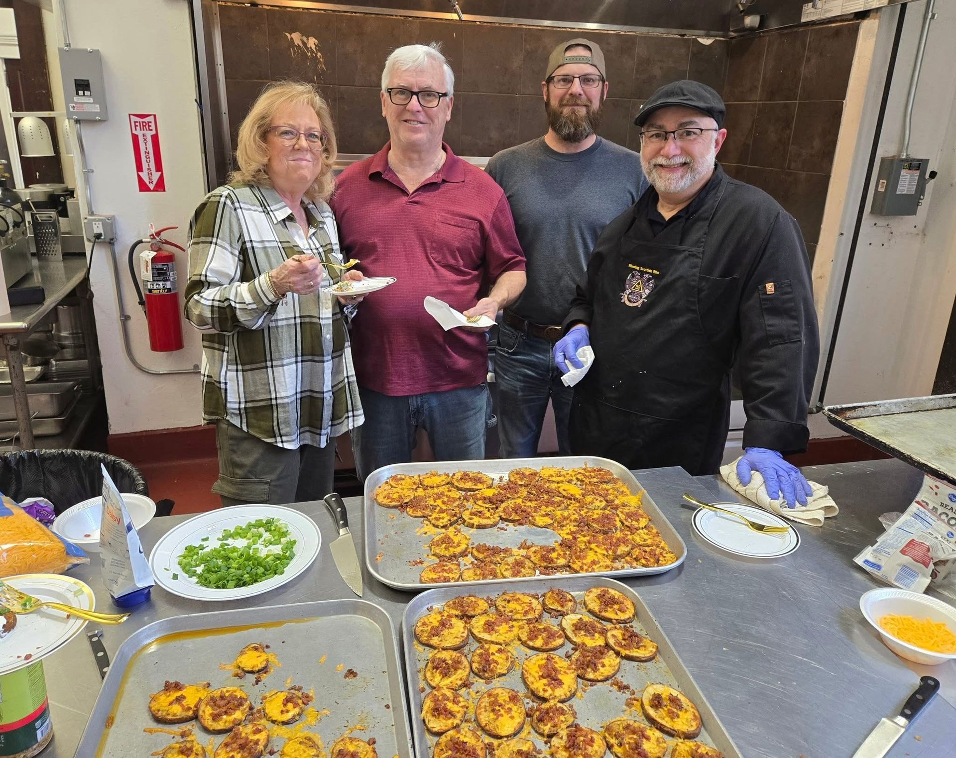 Four people in a kitchen with trays of food. Smiling, they stand near cooked potato snacks.