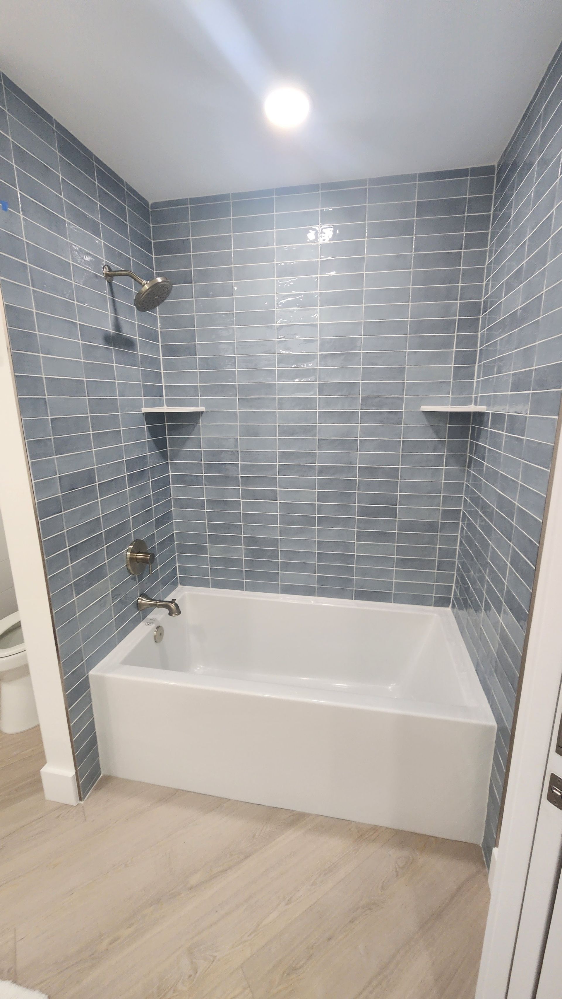 Small tiled bathroom with a white tub, gray wall tiles, and wood-look floor.