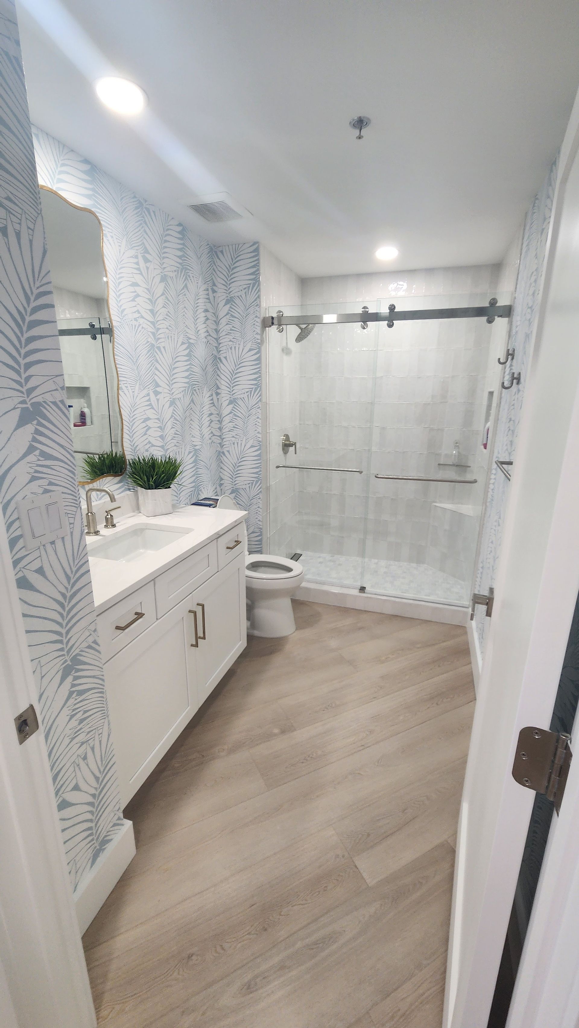 Bright bathroom with white vanity, patterned wallpaper, wood floor, and glass shower door.