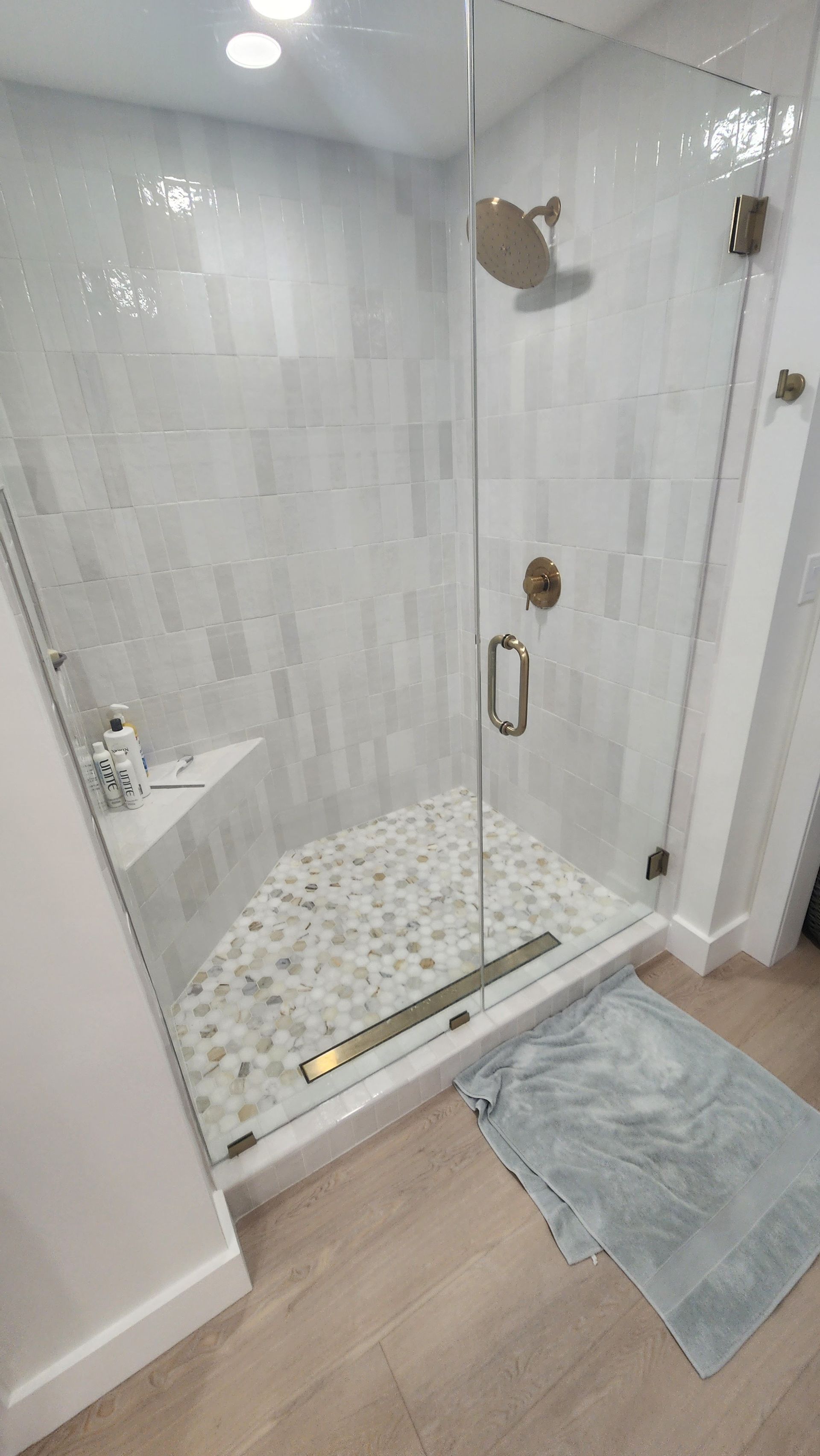 Glass walk-in shower with white tile, chrome door handle, and a blue bath mat on wood-look floor