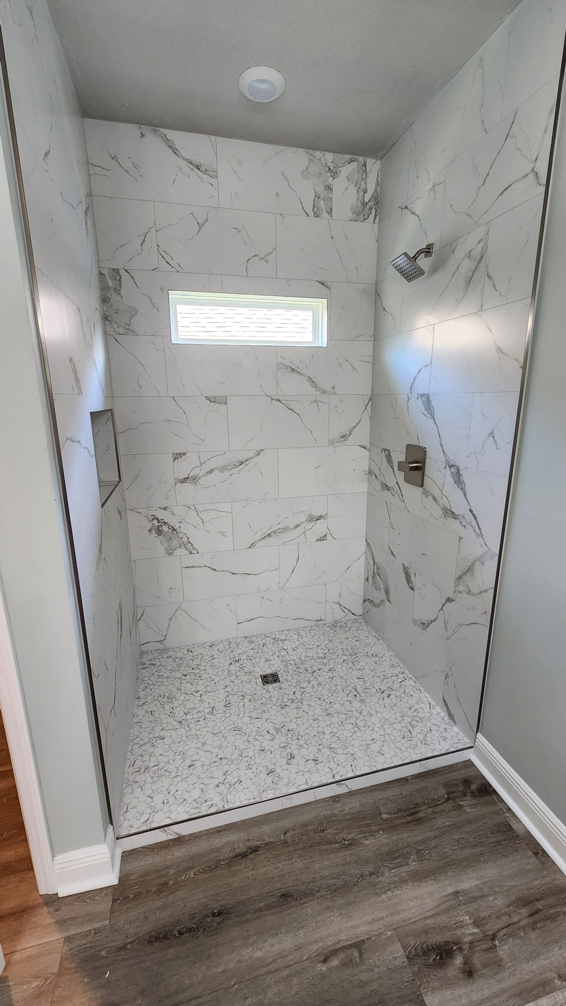 Empty marble-tiled shower with a small window and built-in niche, viewed from a wood-floored bathroom.