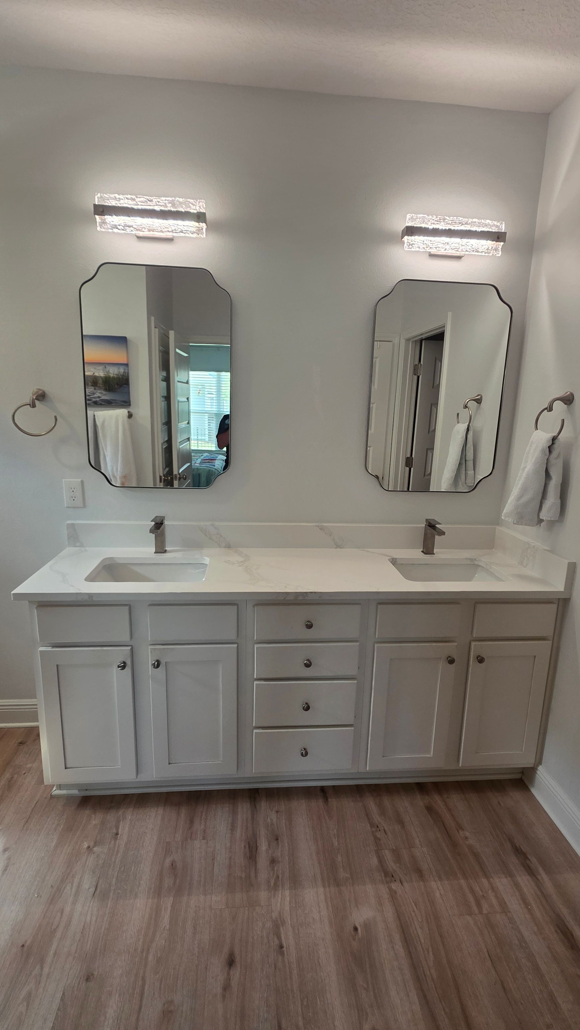 Modern bathroom vanity with double sinks, white cabinets, mirrors, and wall sconces over wood-look flooring