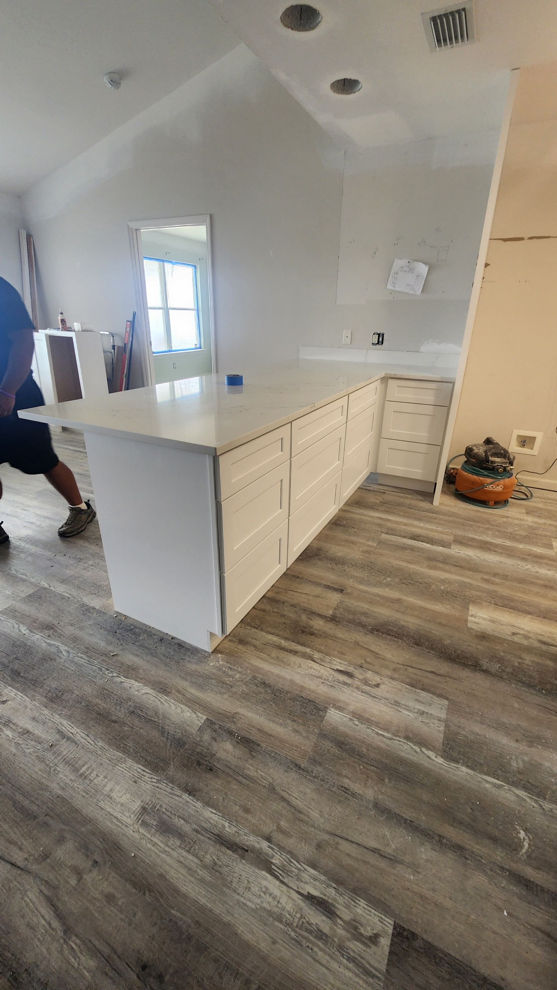 Modern kitchen with white cabinets, gray wood floors, and a central island; person partially visible at left.