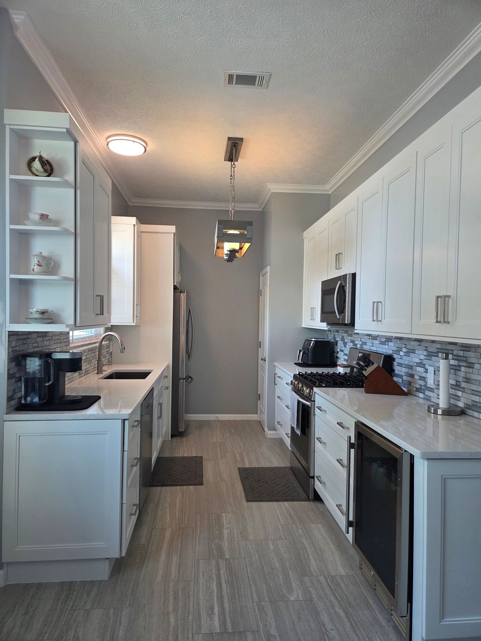 Narrow galley kitchen with white cabinets, gray floors, and stainless steel appliances, lit by ceiling lights.