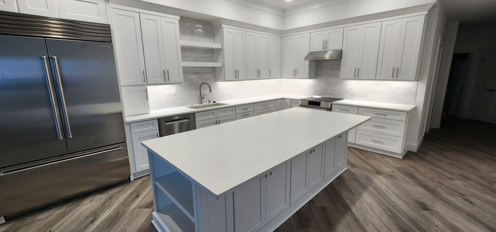 Modern white kitchen with large island, gray cabinets, stainless steel fridge, and wood-look flooring