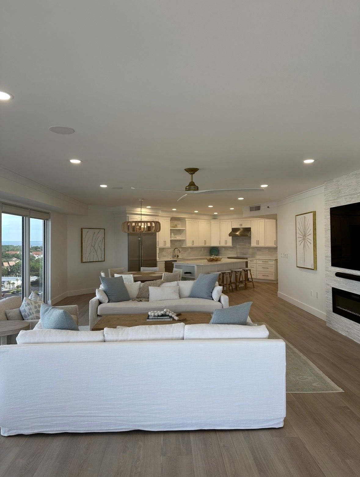 Bright open-concept living room with white sofas, fireplace, and kitchen in the background