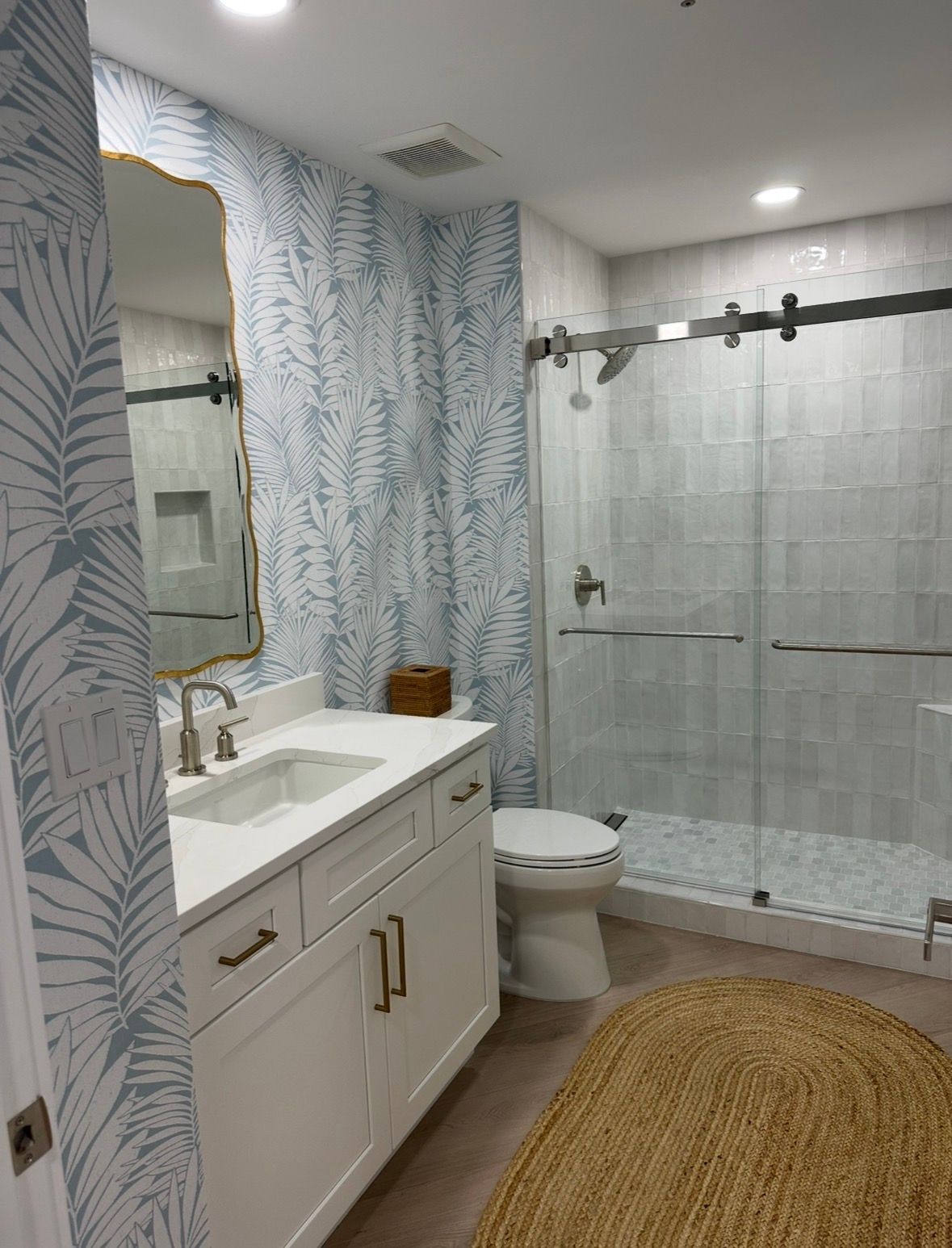 Bright bathroom with white vanity, toilet, glass shower, blue patterned curtain, and woven rug.