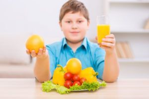 A young boy is sitting at a table holding an orange and a glass of orange juice.