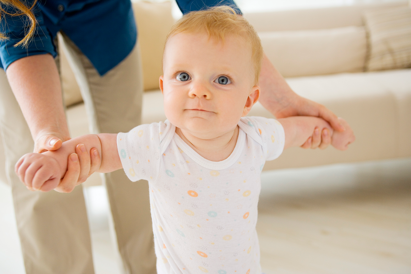 Baby learning to walk, held by a person. Cream-colored sofa in background.