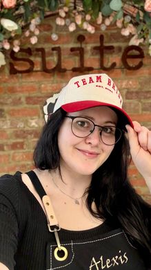 A woman wearing a hat and glasses is standing in front of a brick wall.