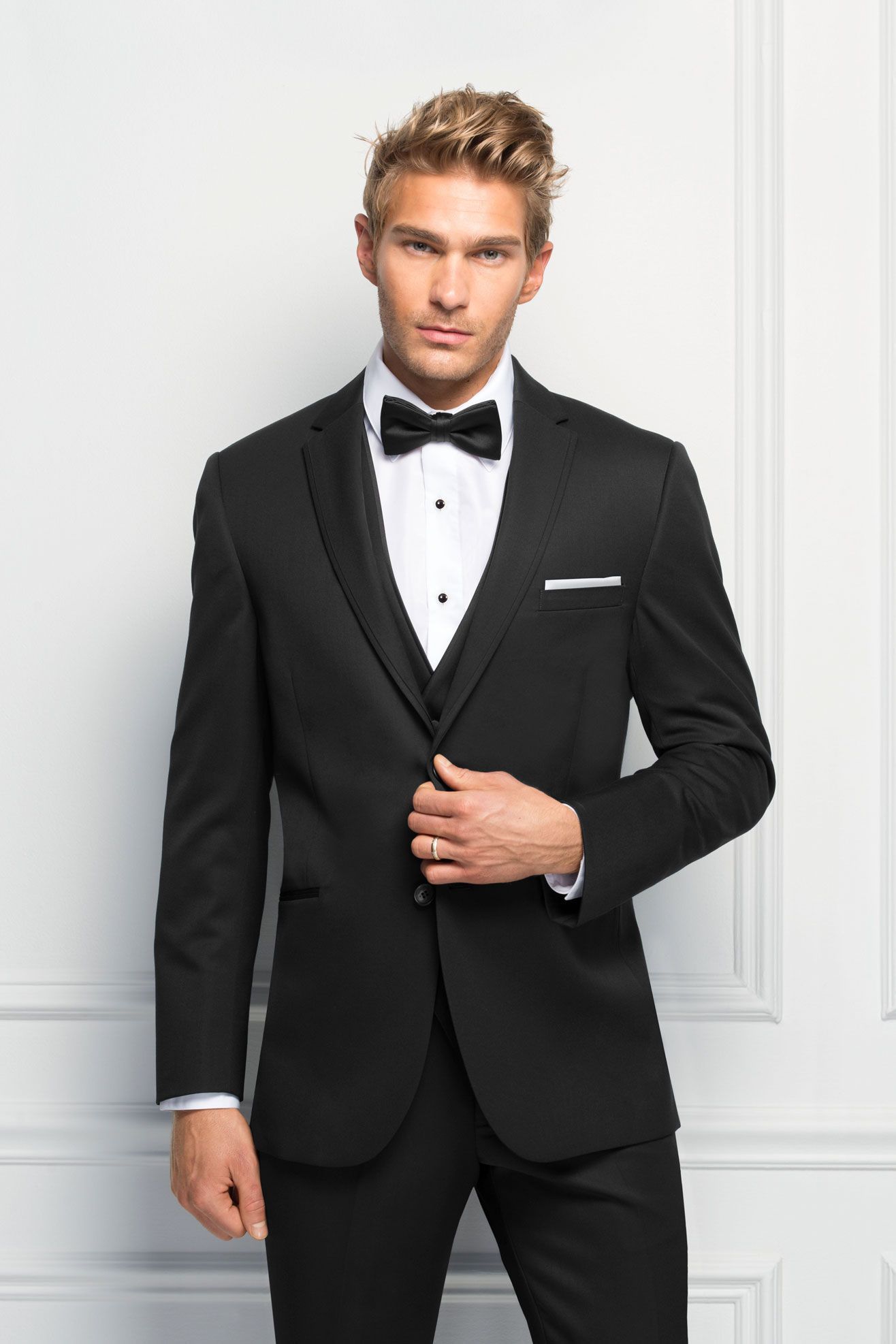 A man in a black tuxedo and bow tie is standing in front of a white wall.