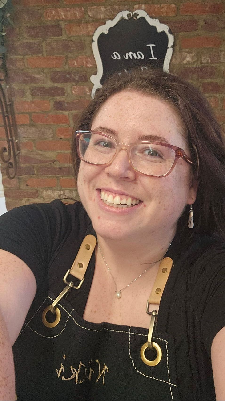 A woman wearing glasses and an apron is smiling in front of a brick wall.