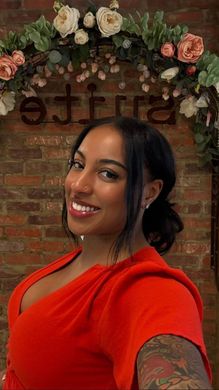 A woman in a red dress is taking a selfie in front of a brick wall.