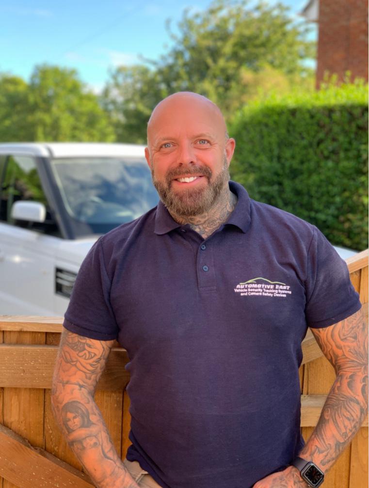 A man with a beard and tattoos is standing next to a wooden fence.