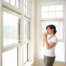 Woman holding a mug, looking out a window. She is in a bright room with white trim, and a field is visible through the glass.