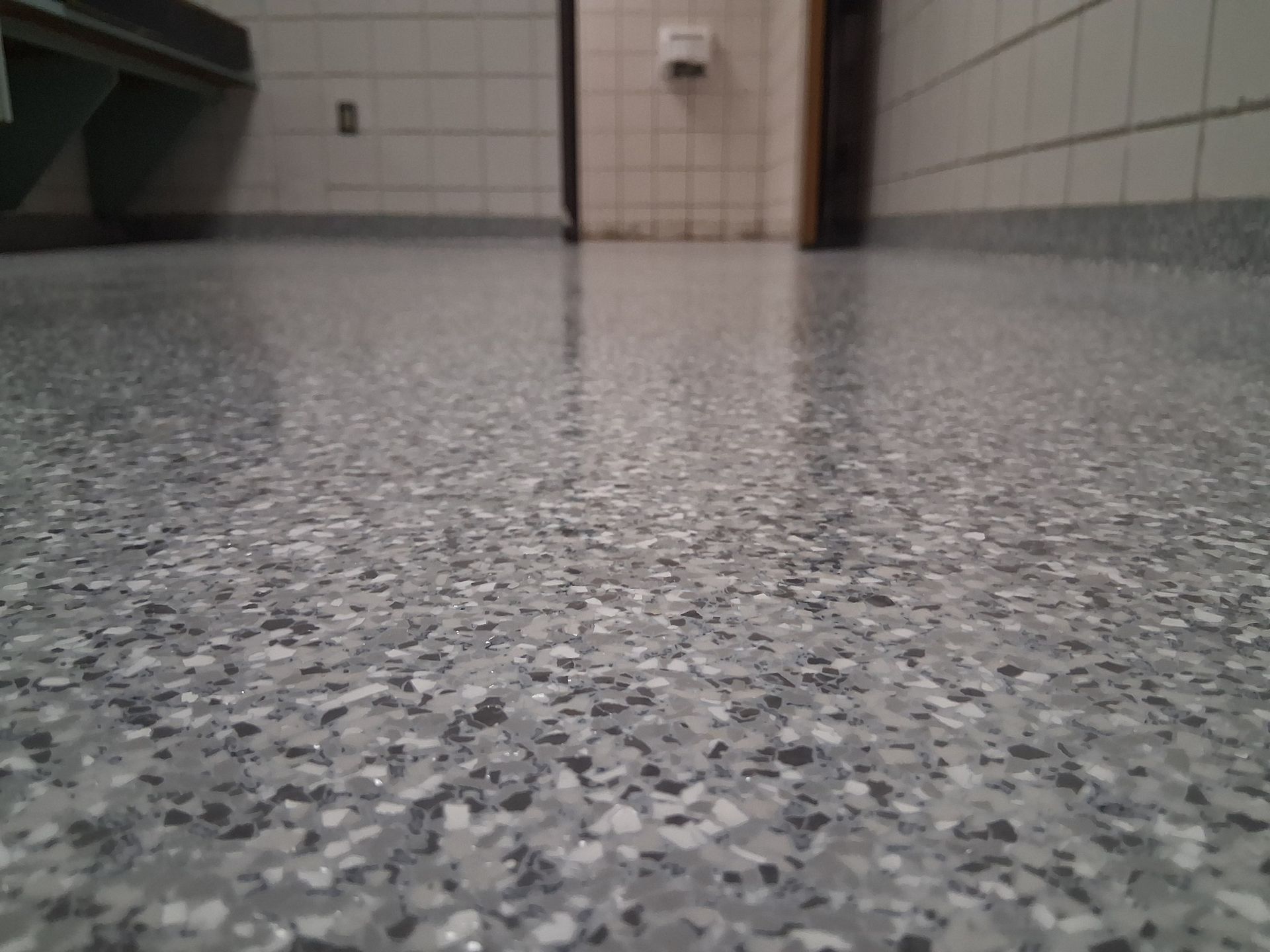 Close-up of a speckled, shiny floor in a public restroom with white tile walls.