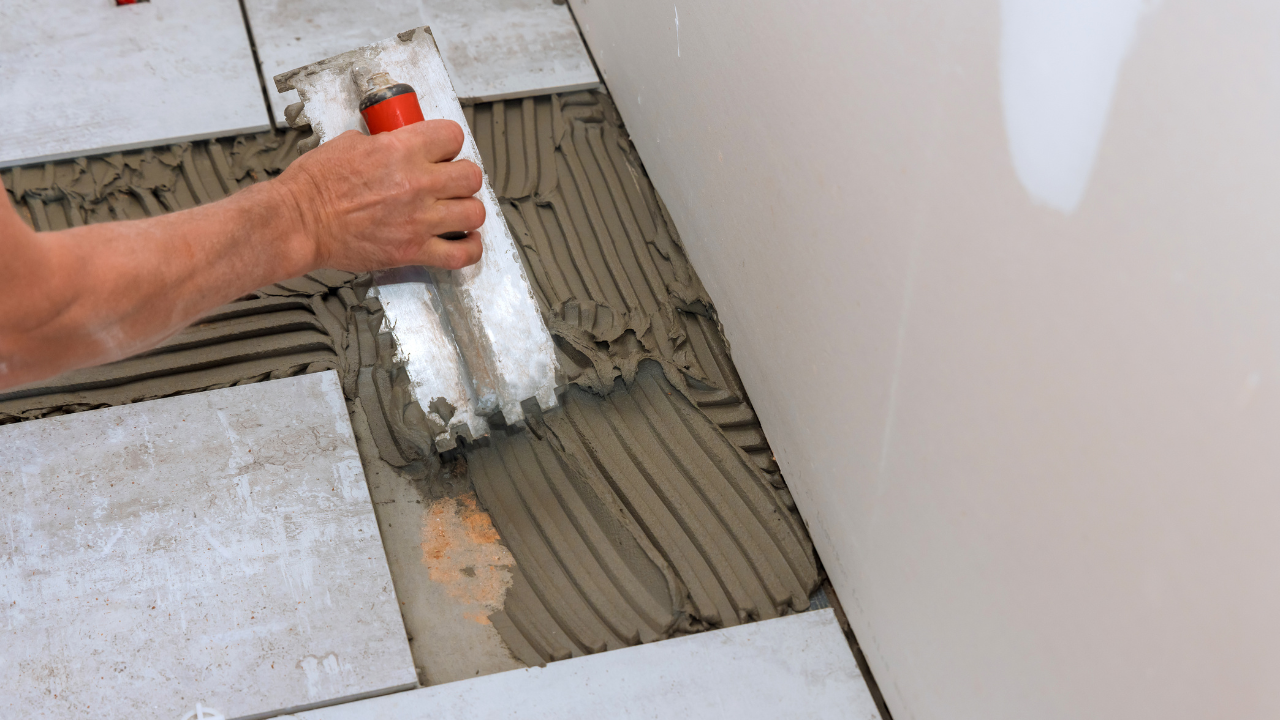 Person using a notched trowel to spread mortar between floor tiles in a room.