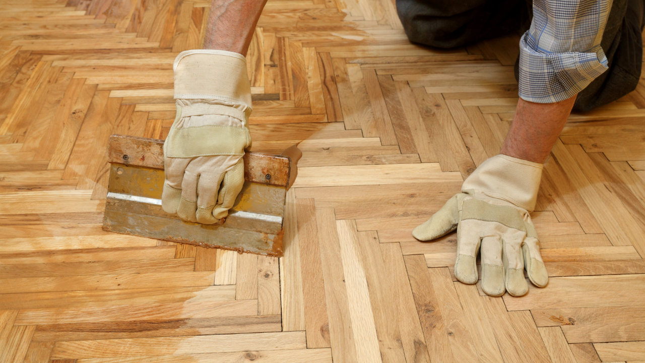 Person with gloved hands applying finish to herringbone wood floor with a trowel.