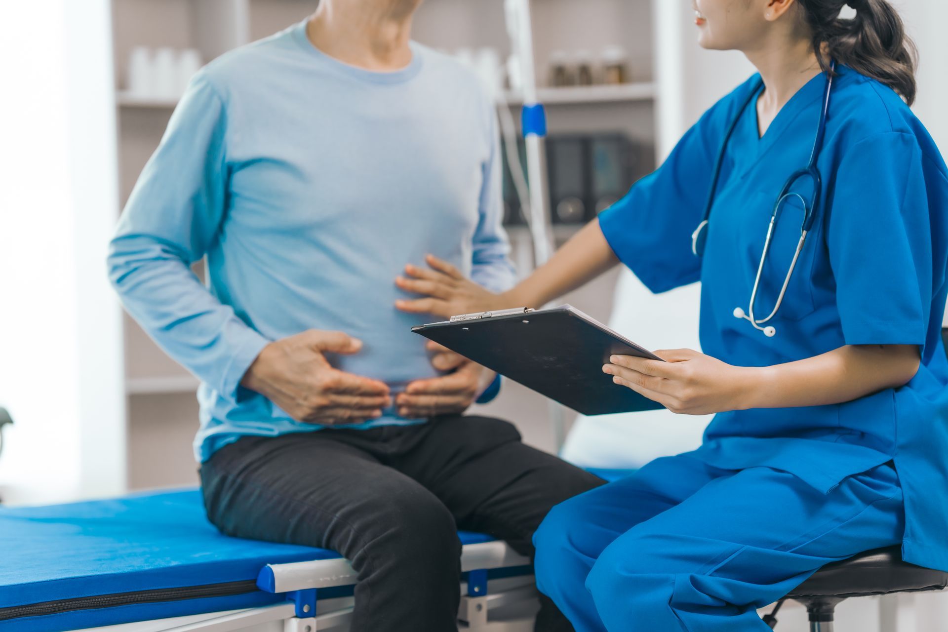 A person sitting on an examination table, holding their stomach while a medical professional in blue scrubs touches their side and holds a clipboard.