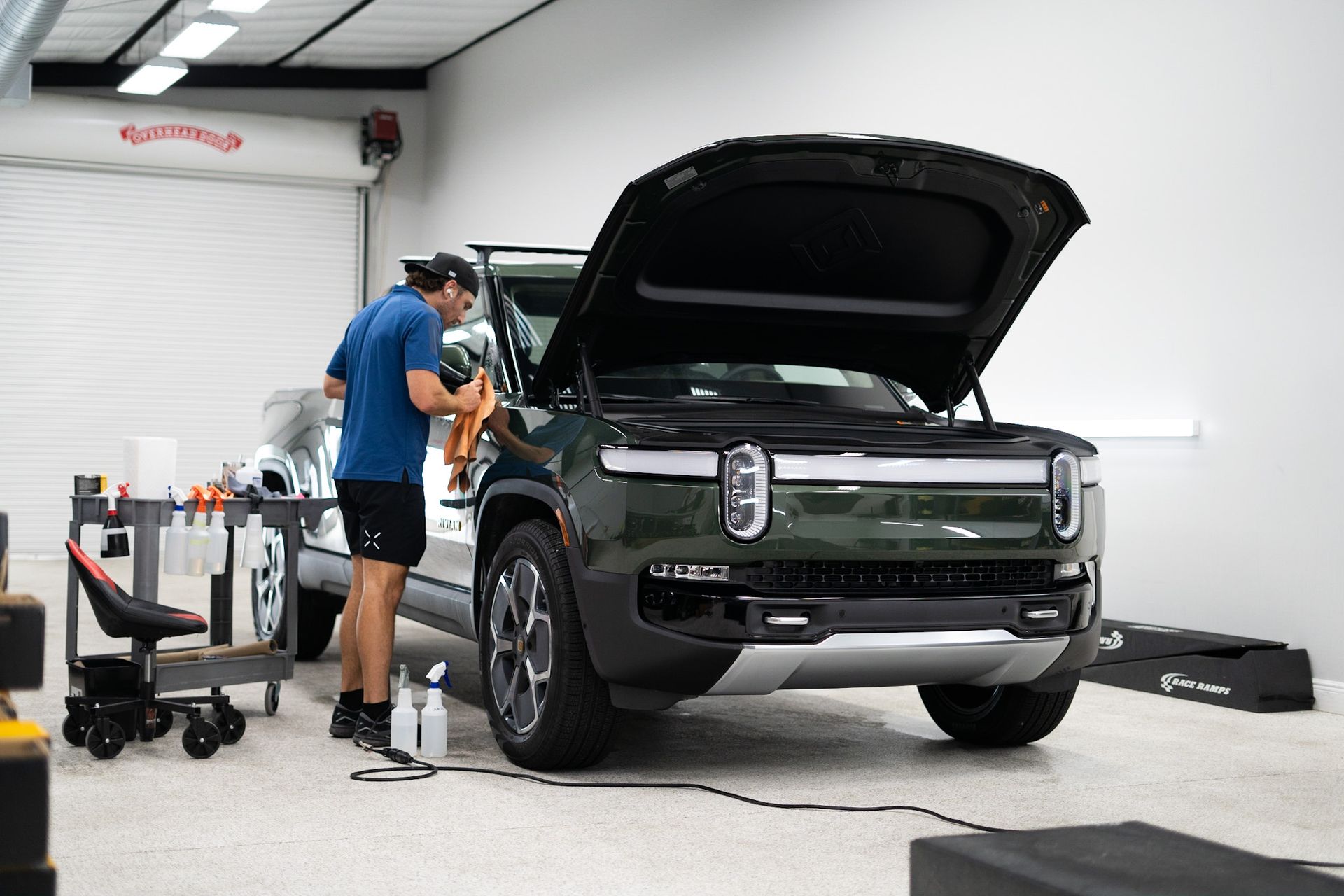 Man working on a dark green Rivian pickup with open hood in a garage.