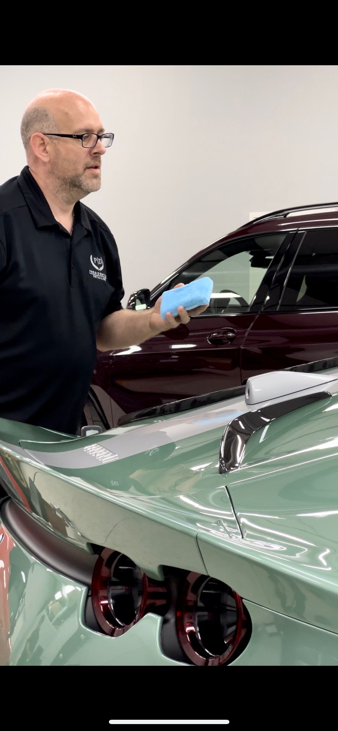 A man with glasses holding a blue sponge near a two-tone car: green and maroon.