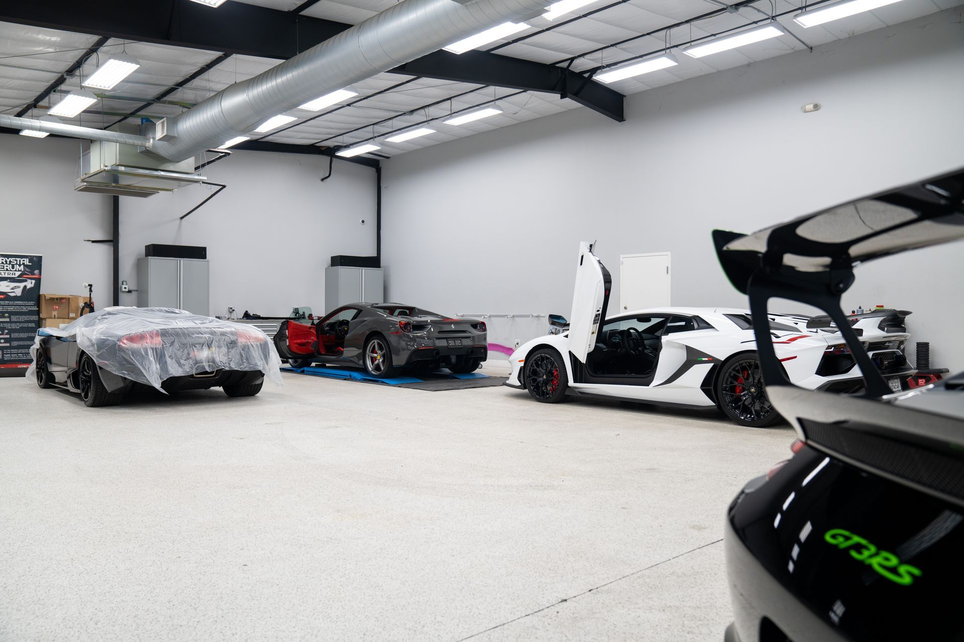 Cars inside a garage, including a Lamborghini with open doors and a covered vehicle; white walls and flooring.