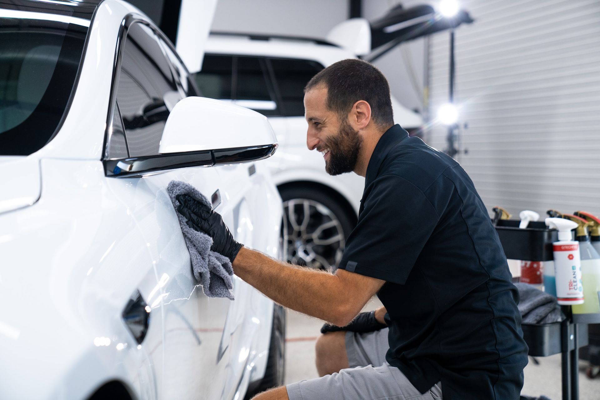 Man detailing a white car, smiling, inside a garage.