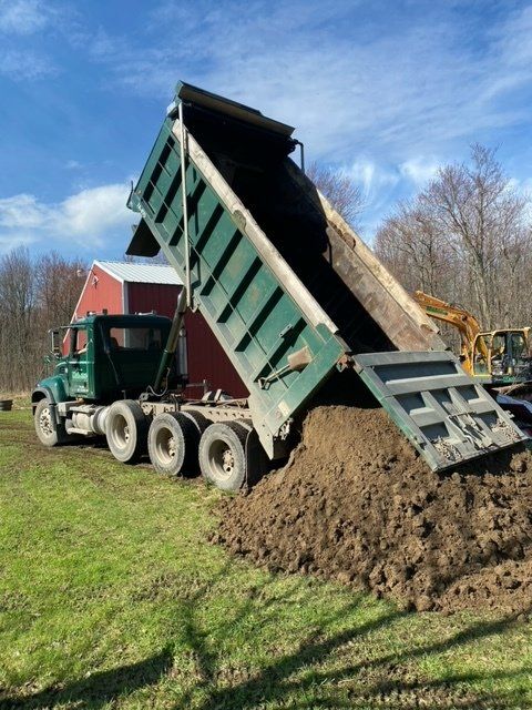 Green dump truck unloading dirt onto a grassy area with a red barn in the background.