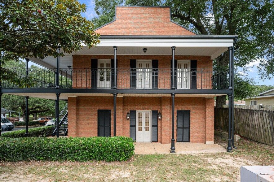 Two-story brick building with black balconies and shutters, surrounded by trees and a hedge.