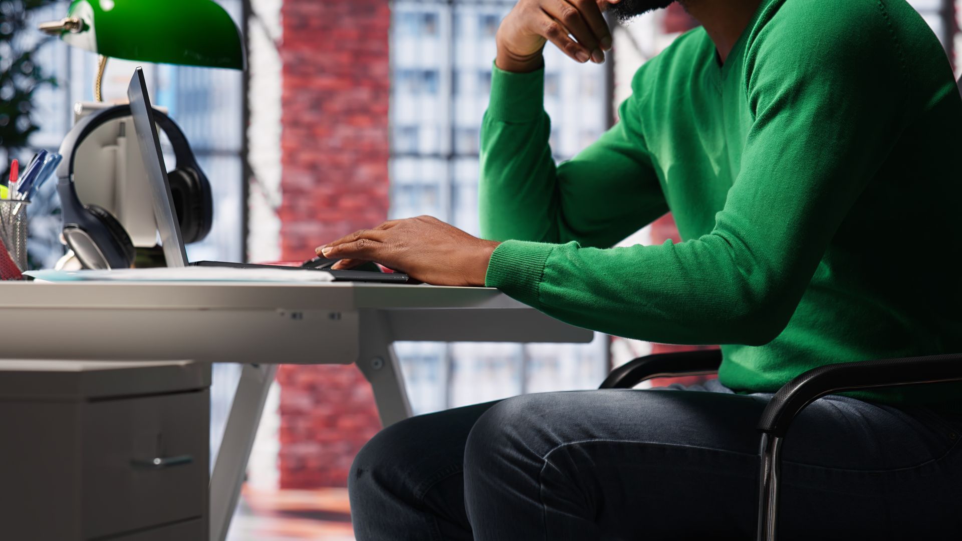 A person wearing a green sweater sits at a desk, typing on a laptop in a bright, modern office.