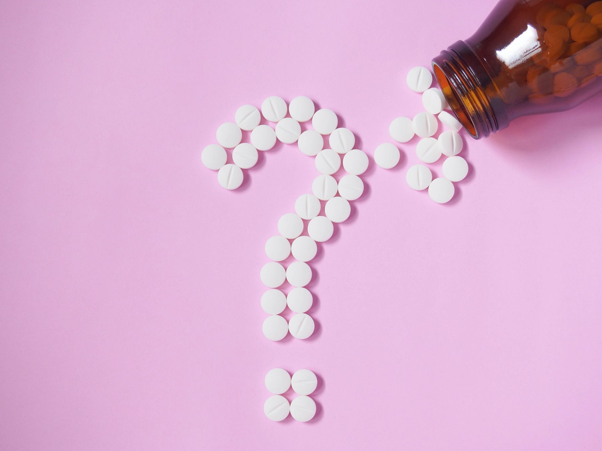 Pills arranged in a question mark shape spilling from a brown bottle on pink background.
