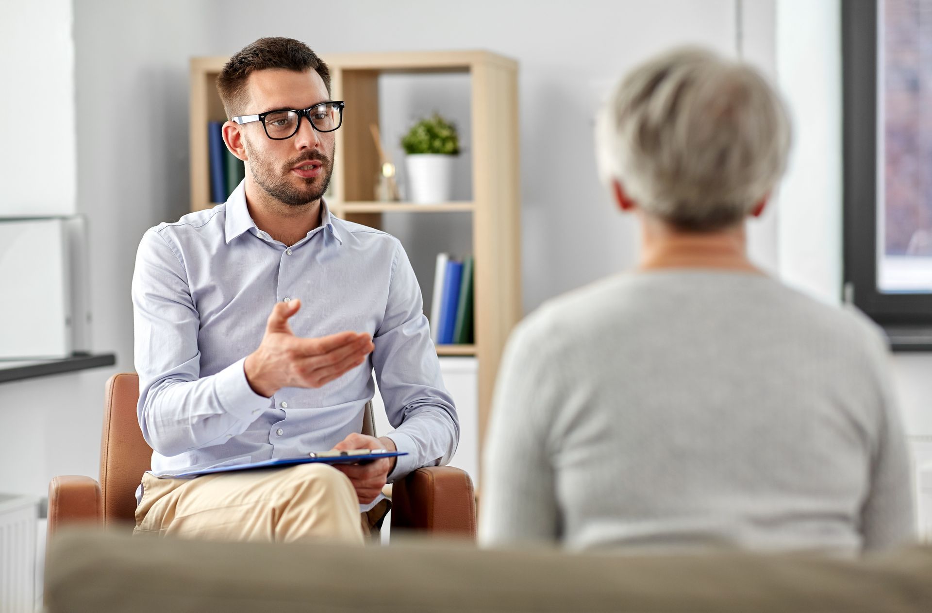 Therapist and patient in consultation. Man gestures, holding clipboard. Neutral background, bookshelves.