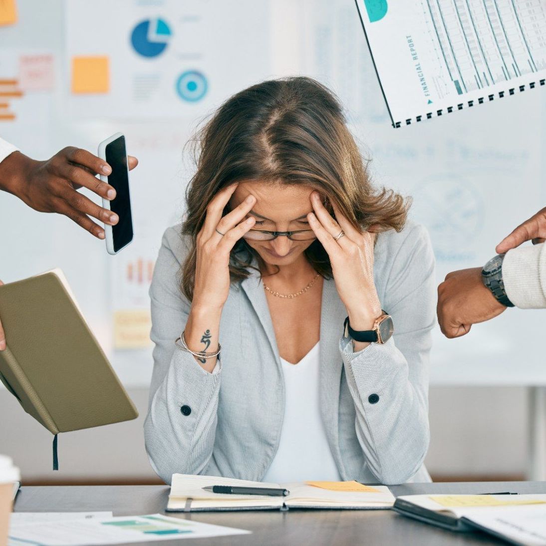 A woman is sitting at a desk with her hands on her head surrounded by people.