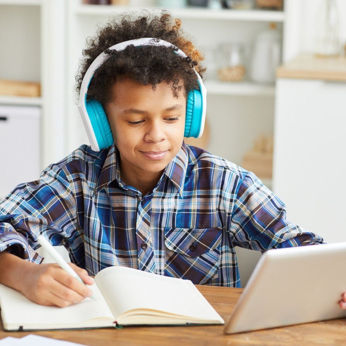 A young boy wearing headphones is writing in a notebook while using a tablet computer.