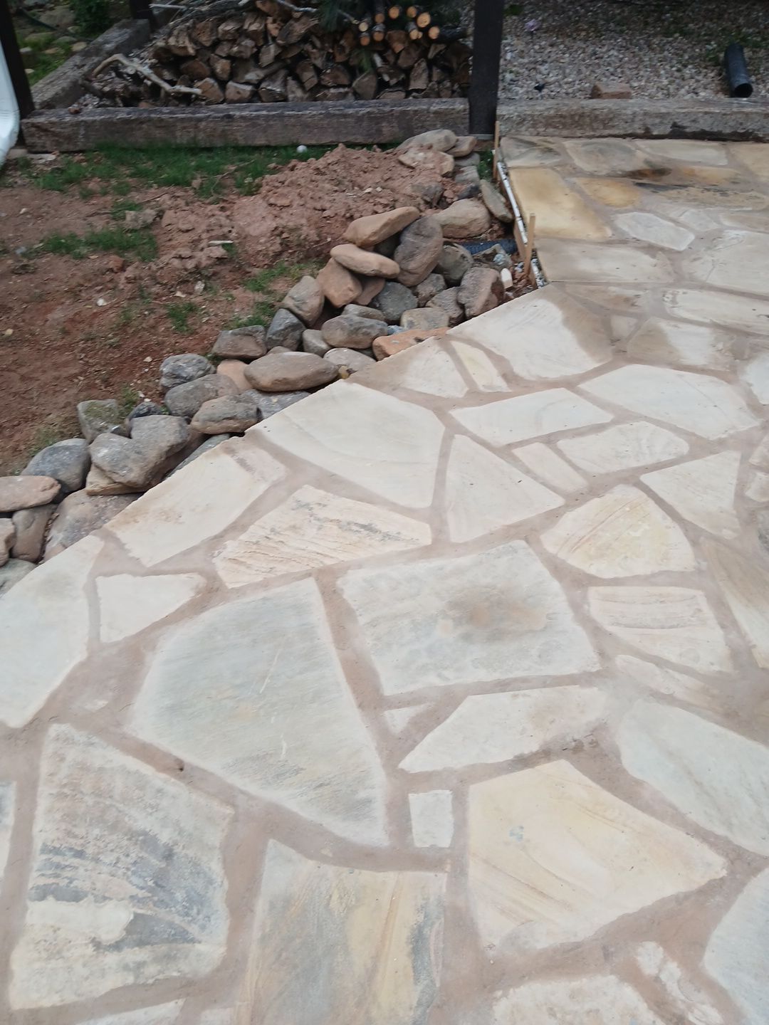 Flagstone patio with gray mortar, bordered by rocks and dirt. Firewood in the background.