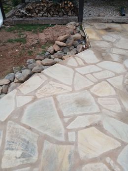 Flagstone patio with gray mortar, bordered by rocks and dirt. Firewood in the background.