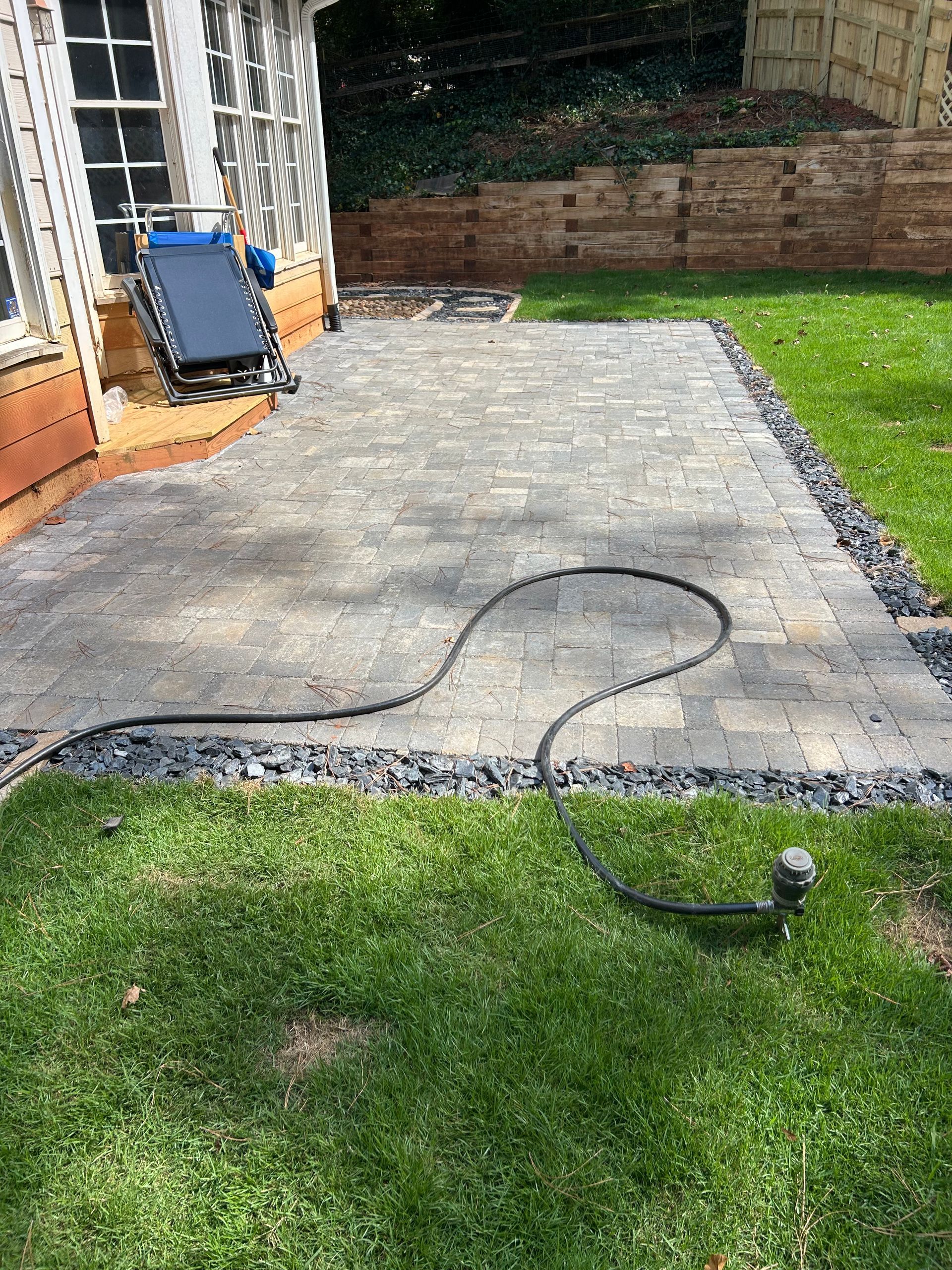 Flagstone patio with gray mortar, bordered by rocks and dirt. Firewood in the background.