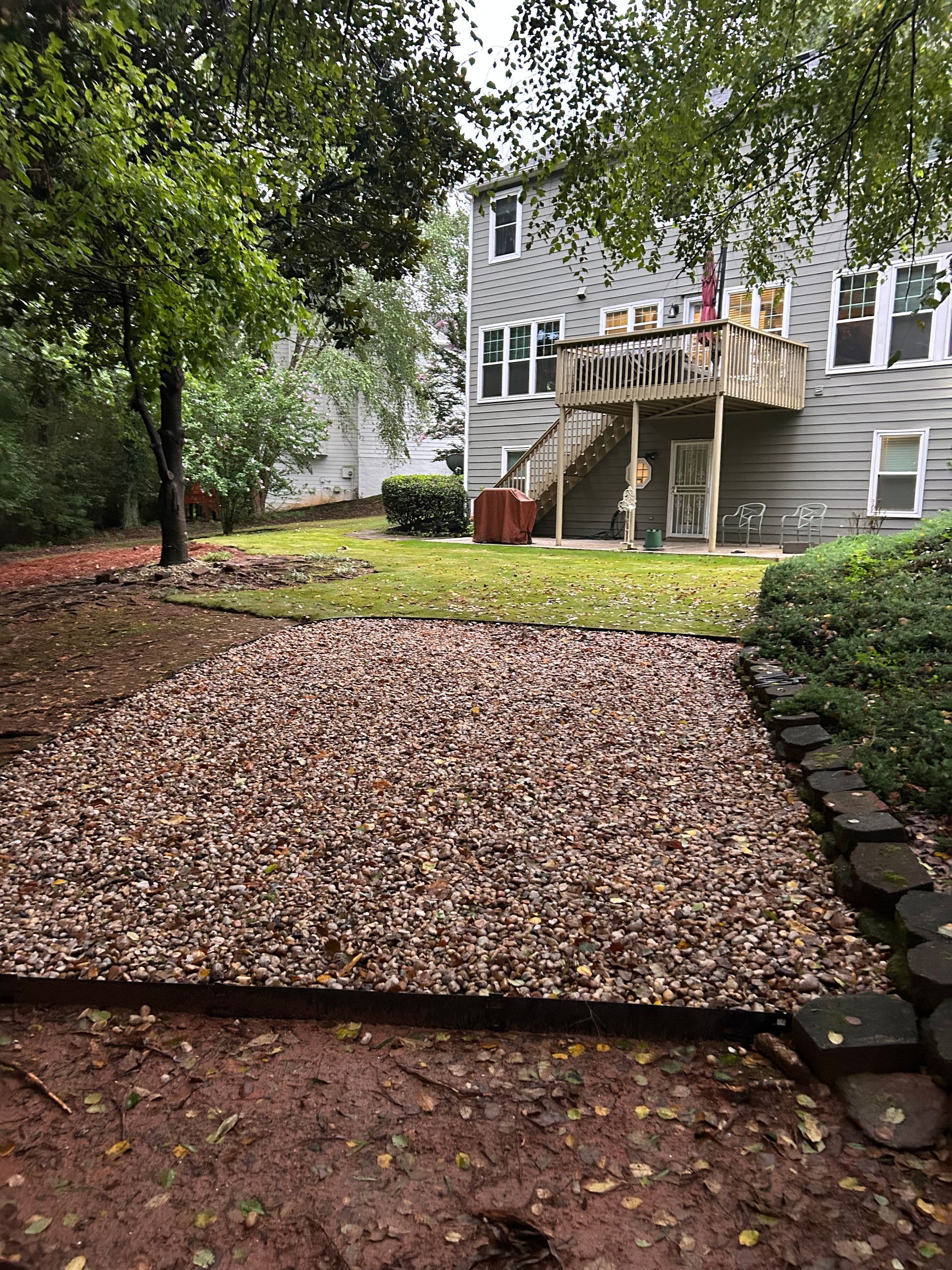 Backyard with gravel area bordered by wood, grass, and a house with a deck.