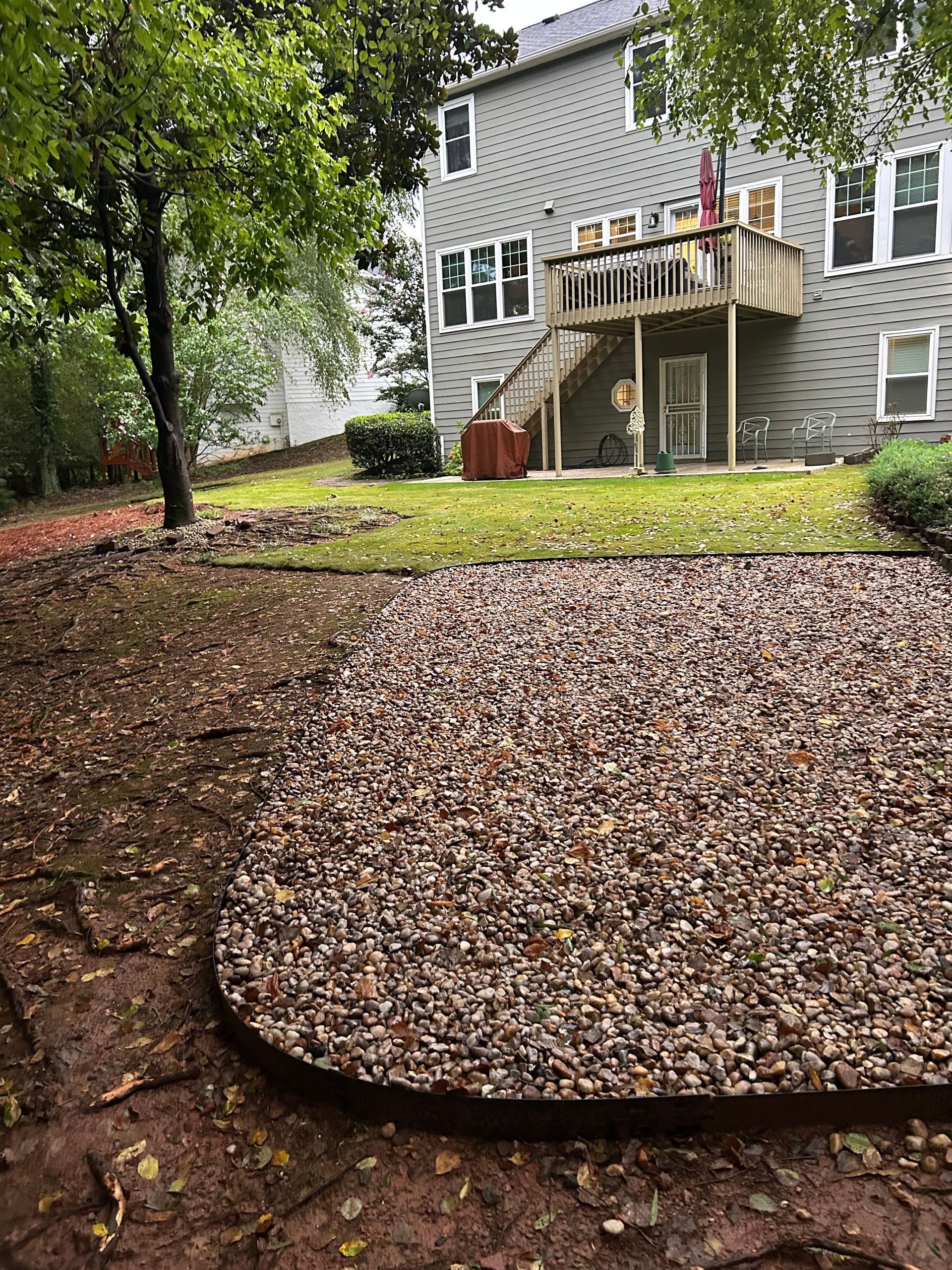 Backyard with gravel patio, grass, and a two-story house with a wooden deck and stairs.