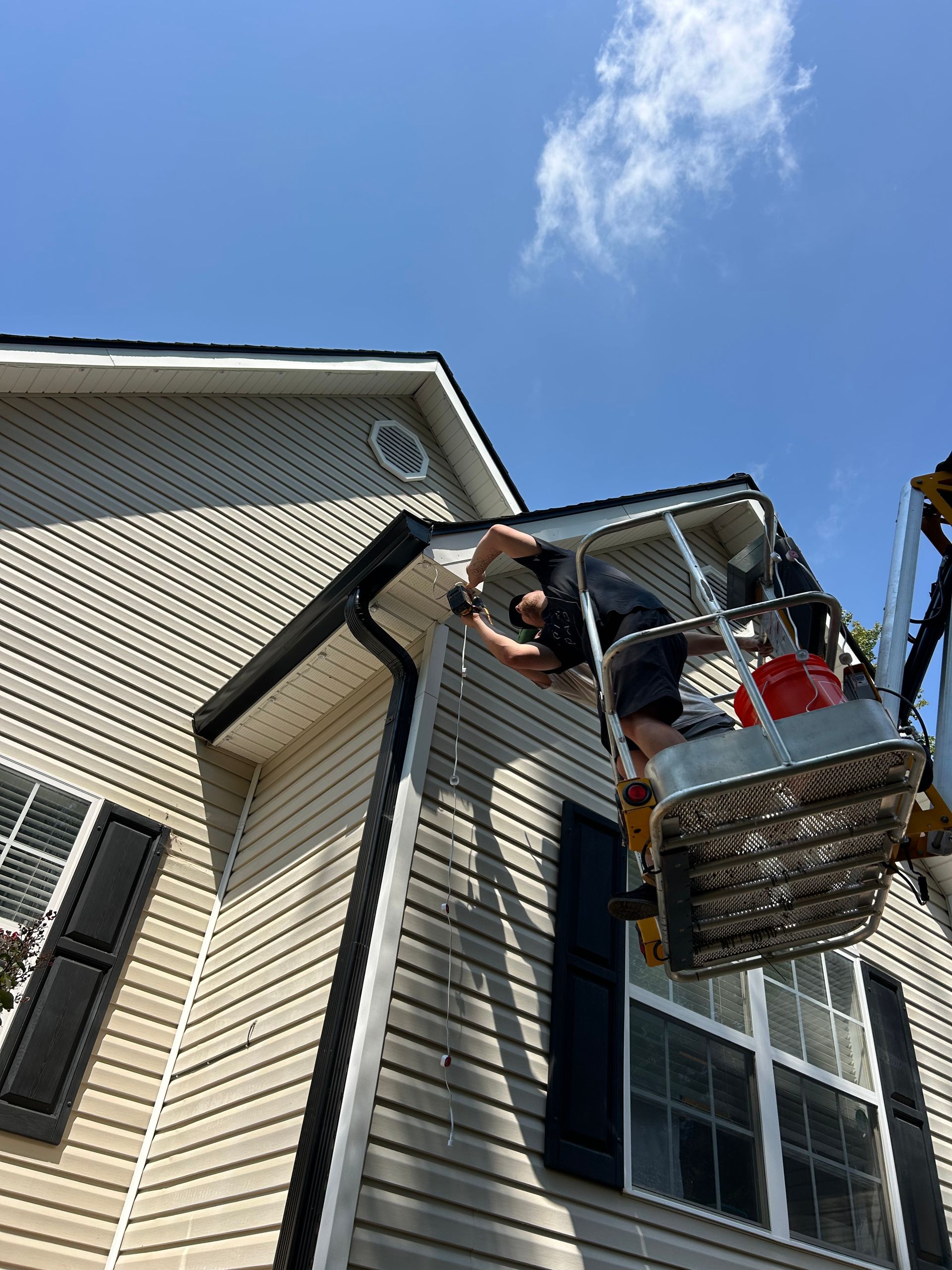 Person in lift basket installing gutter on a beige house, blue sky background.