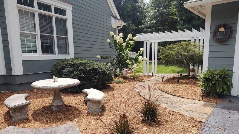 A garden with a stone table and benches, a pergola, and a pathway with greenery and brown mulch.