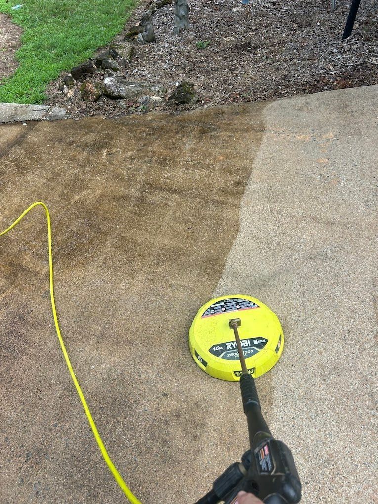 Power washer cleaning a concrete surface; yellow cleaner head, tan concrete, green grass.