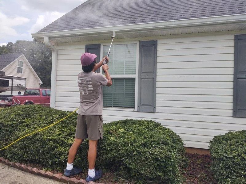 Person power washing a house gutter, spraying water. The house has white siding, gray shutters, and a bush.